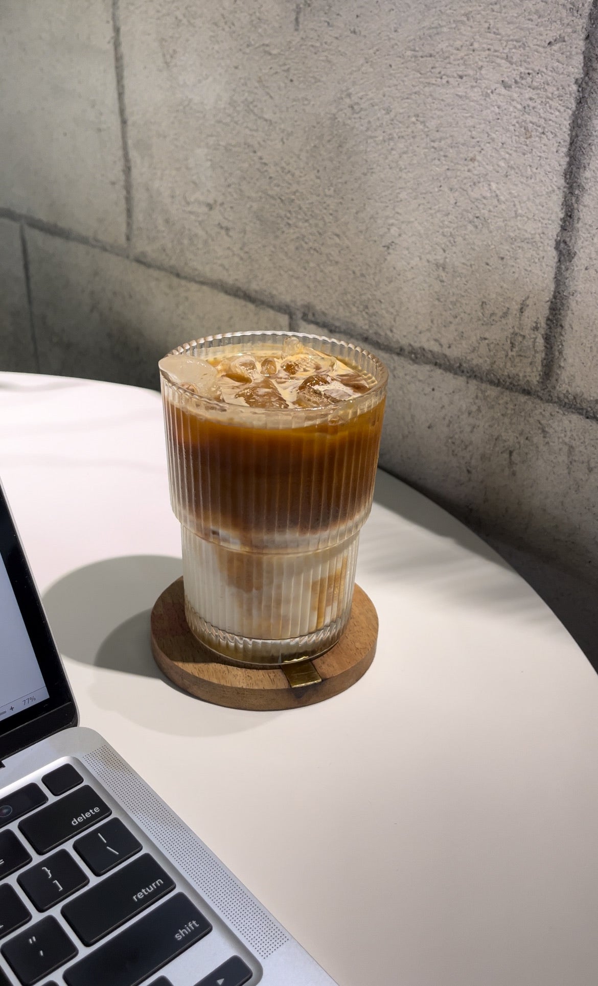 Glass of iced coffee on a coaster next to a laptop on a white surface with a gray textured wall background